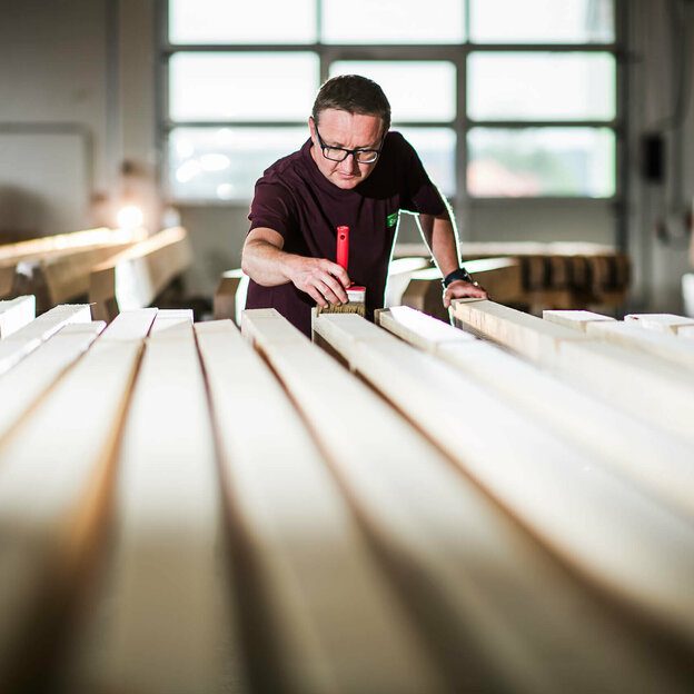 Mitarbeiter streicht Holz mit einem roten Pinsel in der Werkstatt